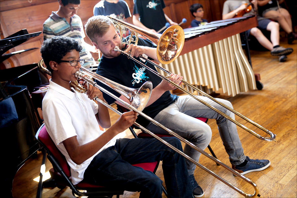 A black student playing the trombone alongside a white male teacher, also playing the trombone, with other students in the background.